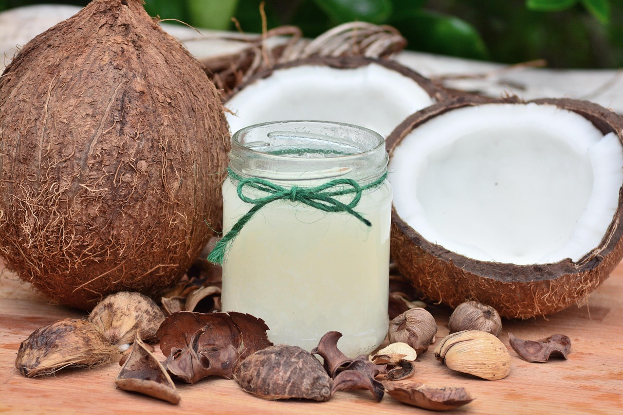 Coco fresco inteiro e partido ao meio, mostrando a polpa branca, ao lado de um potinho de óleo de coco