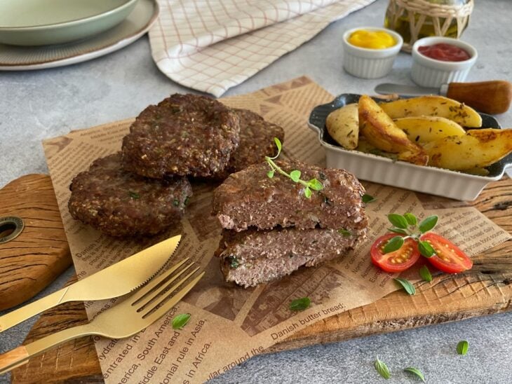 Hambúrgueres fit servidos numa tábua de madeira com batatas assadas, tomate-cereja e talheres dourados