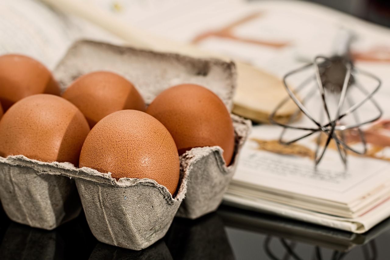 Ovos numa caixa de papelão ao lado de um batedor de arame, sobre um livro de receitas