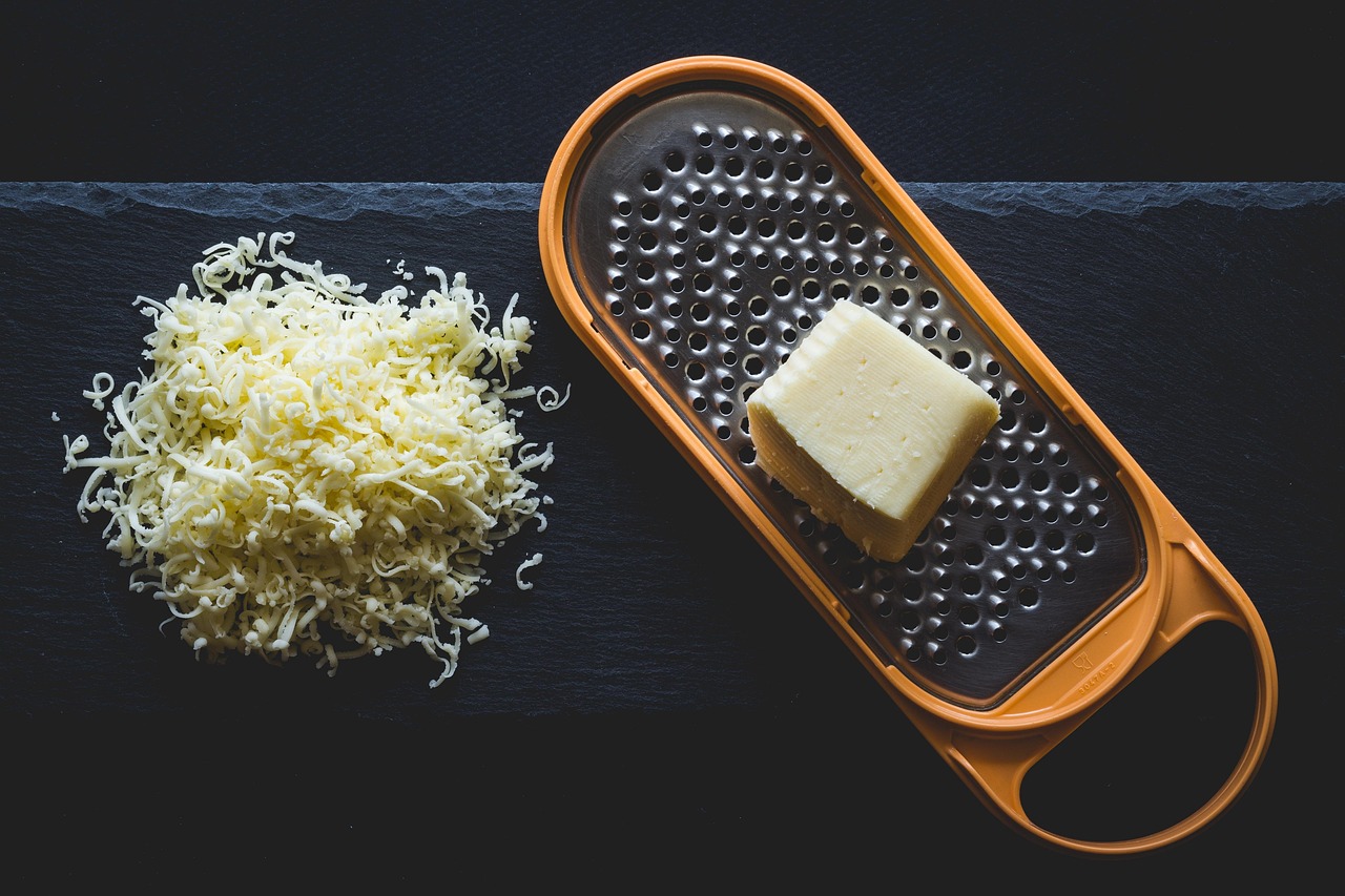 Queijo ralado ao lado de um cubo de queijo sobre um ralador, em uma bancada escura